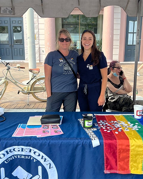 Two individuals stand together behind a table draped with tablecloths and a Pride flag