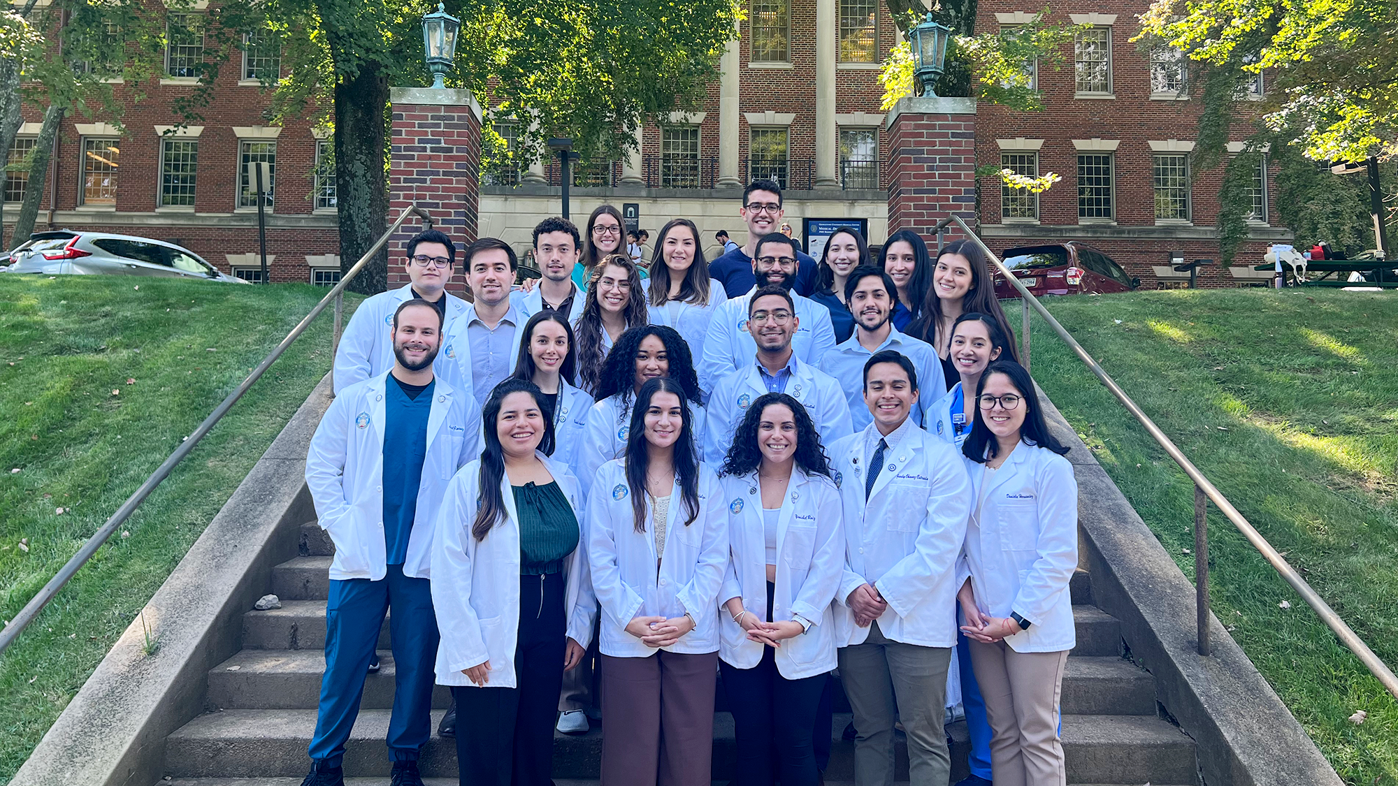A group of medical students in white coats stand together on the steps in front of the Med-Dent Building on Georgetown's campus