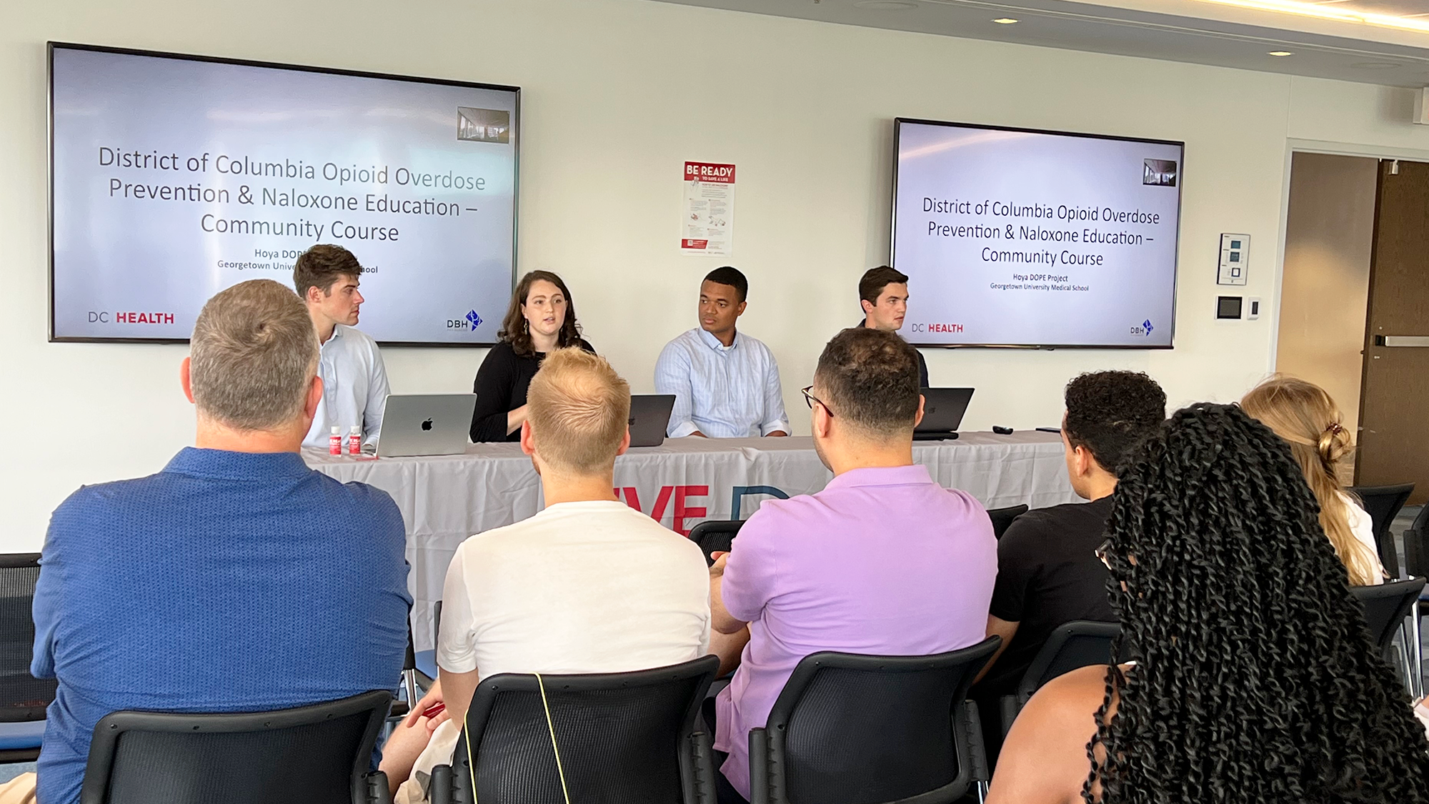 A table of presenters speaks to an audience seated before two large screens