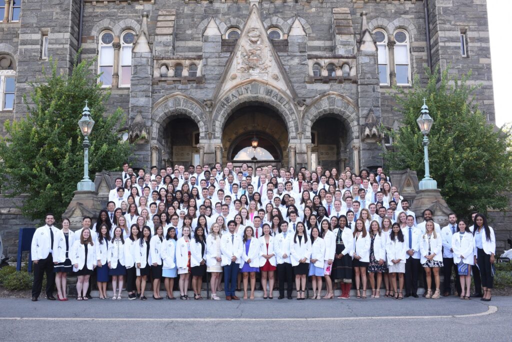 A large group of medical students in white coats stand on the steps of Healy Hall