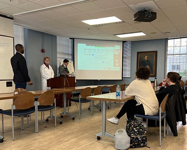 Three individuals stand in a room presenting to several people seated before them at a table