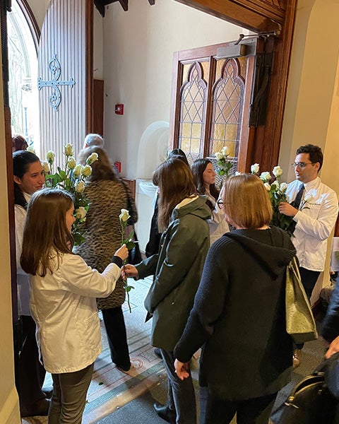 Med students wearing white coats hand flowers to people walking into a chapel