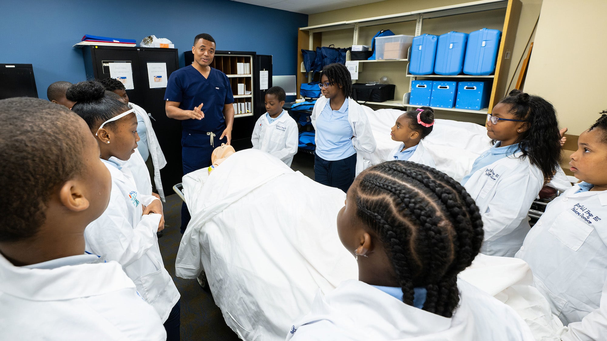 A medical student in scrubs speaks with young students in the simulation lab