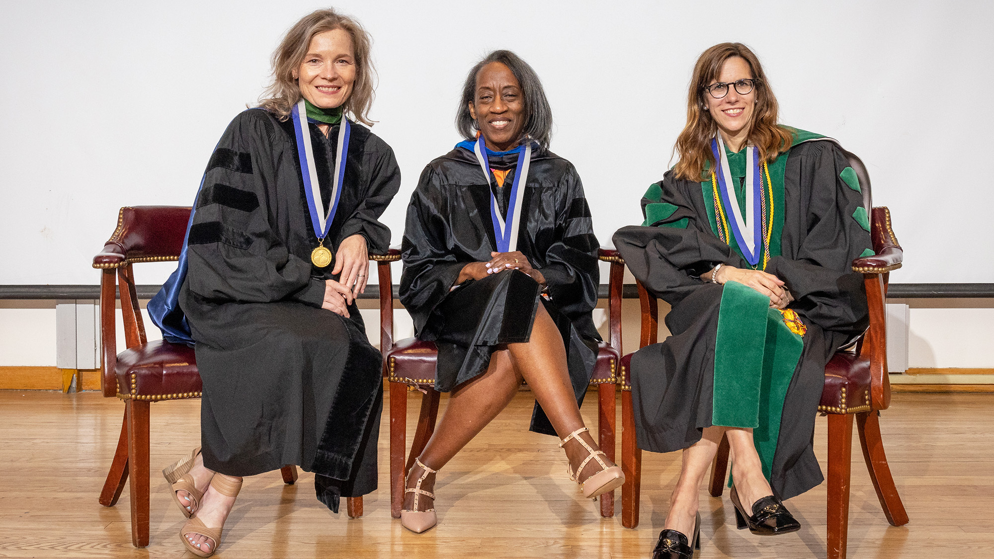 Amy Burke, MD, MPH; Donna Cameron, PhD, MPH; and Mary Furlong, MD, sit in chairs on a stage wearing their academic regalia and MAGIS medallions