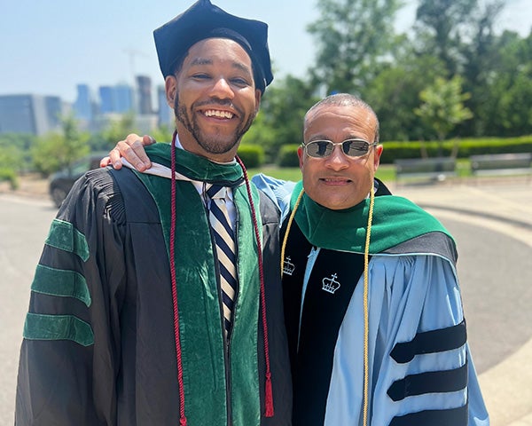 Malcolm Meredith and Dean Lee Jones stand side by side outdoors on campus dressed in academic regalia