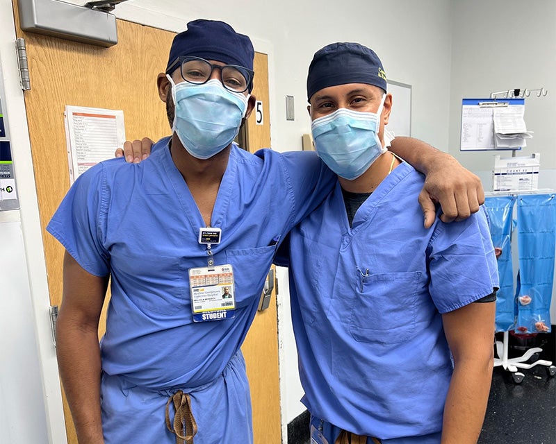 Malcolm Meredith stands with Dr. Aguirre in the OR, both wearing surgical scrubs, hats and masks