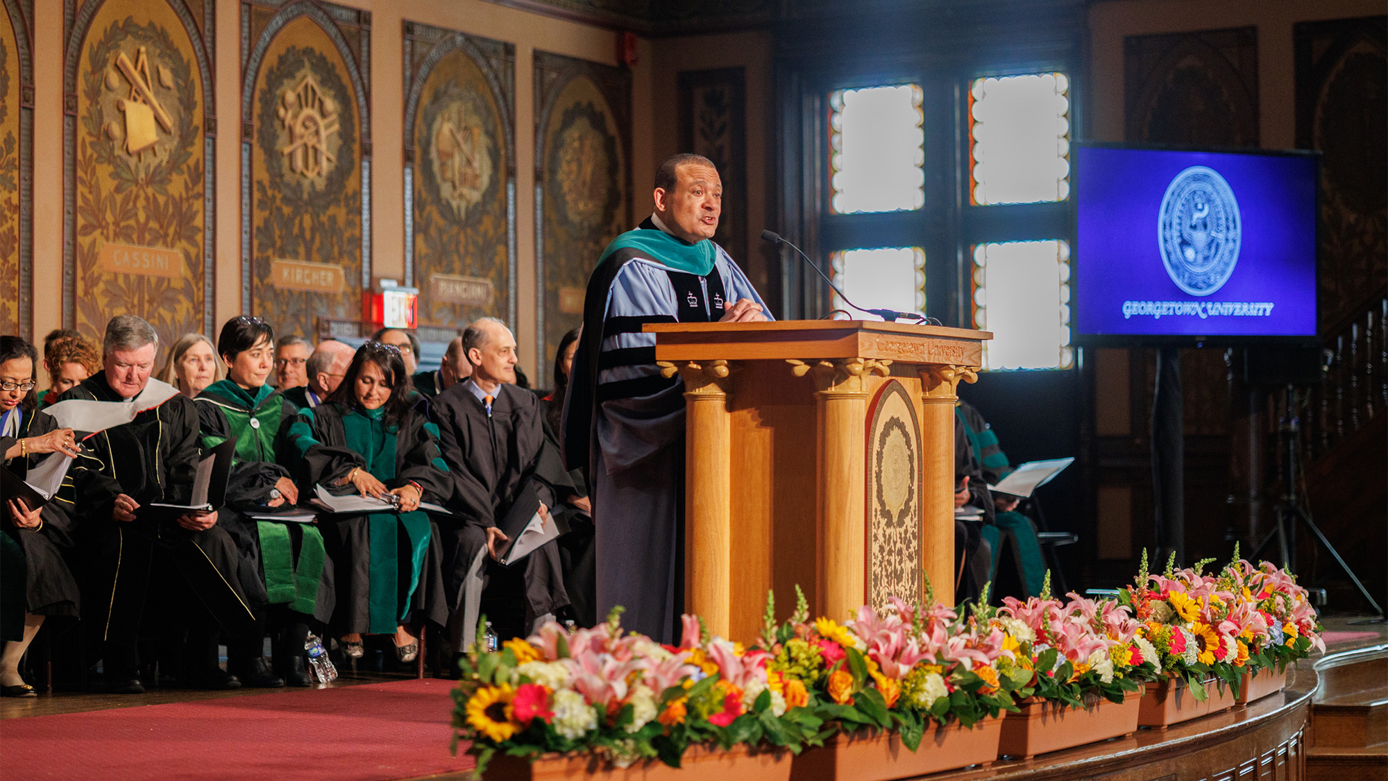 Dean Jones speaks at a podium on the stage of Gaston Hall with SOM faculty seated behind him