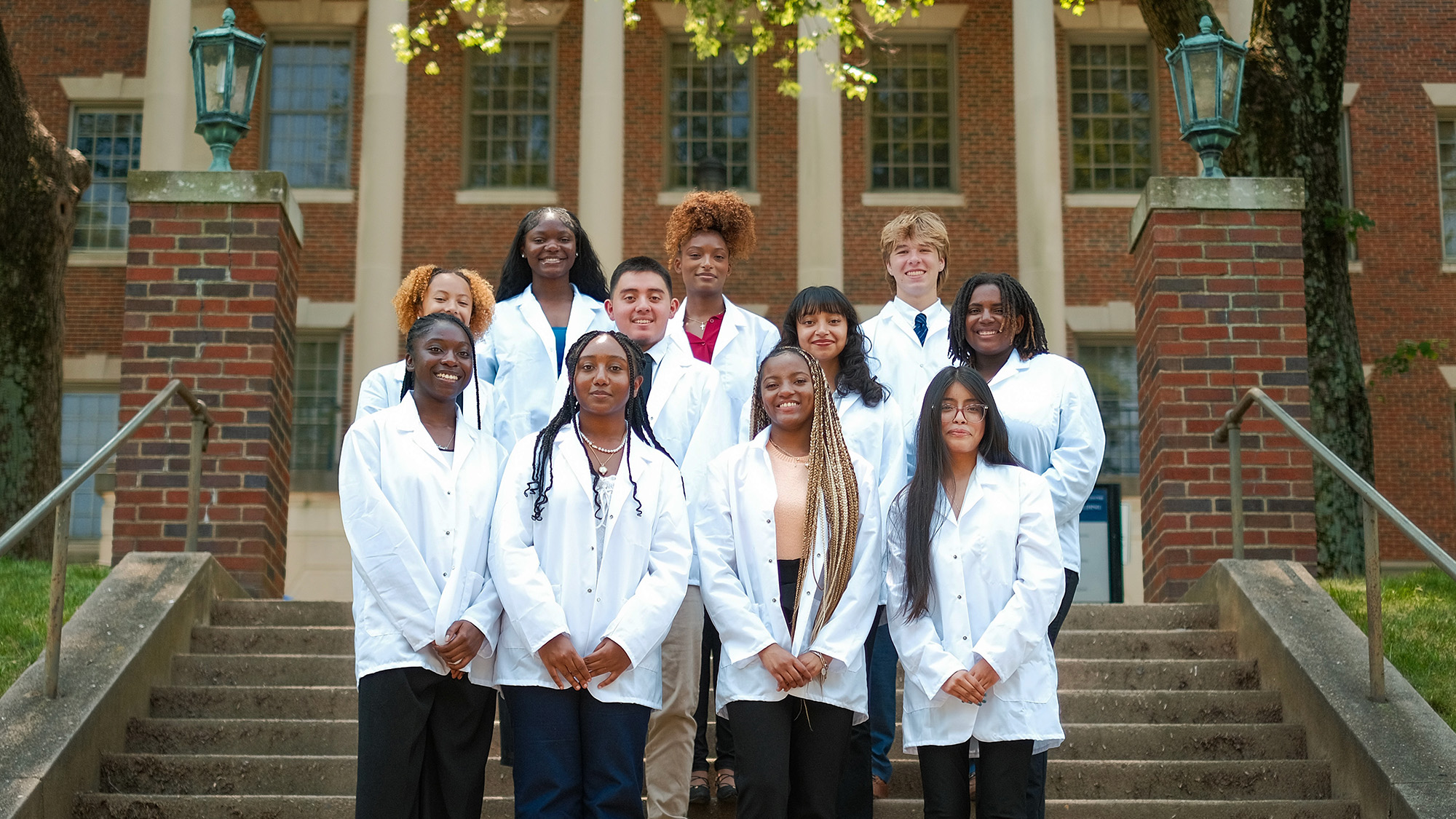 The eight students who were GEP scholars stand on steps in front of the Med-Dent Building on campus wearing white coats