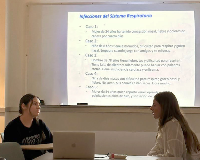 Two students speak to each other while behind them are several case studies projected on a screen