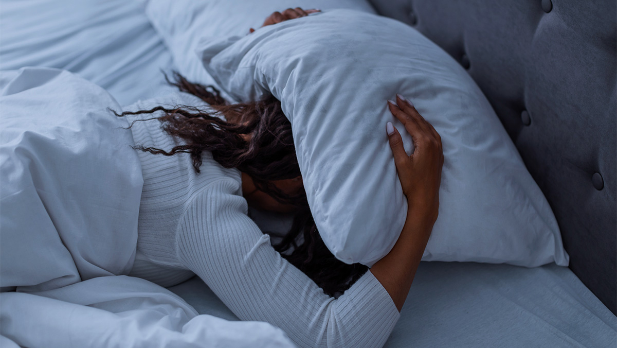A woman lies in bed with her head covered with a pillow