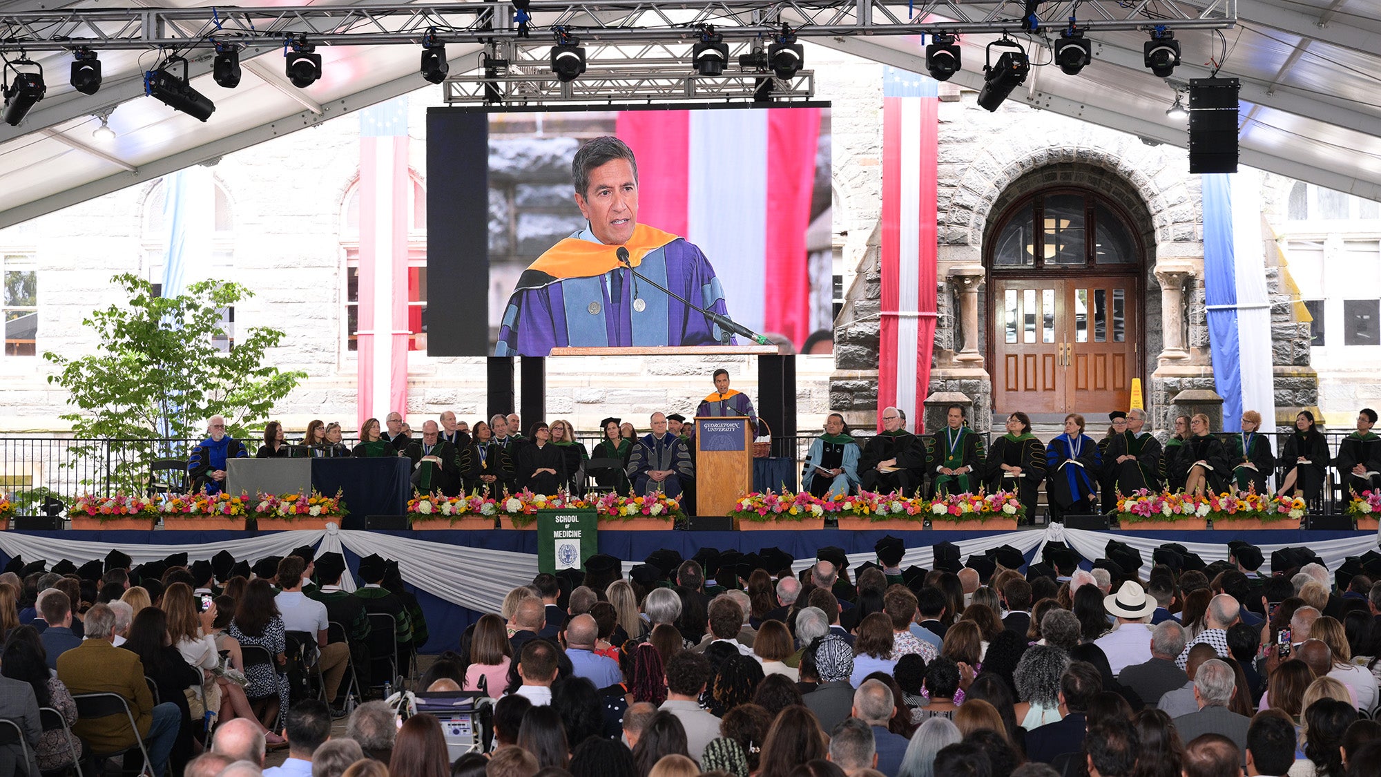 A long view of the stage at commencement with all the faculty seated in a row on either side of the podium where Sanjay Gupta speaks before a large audience