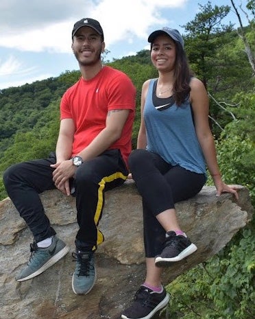 Michael Sobalvarro and Melanie Diaz sit side by side on a rock outcrop in a hilly forested area