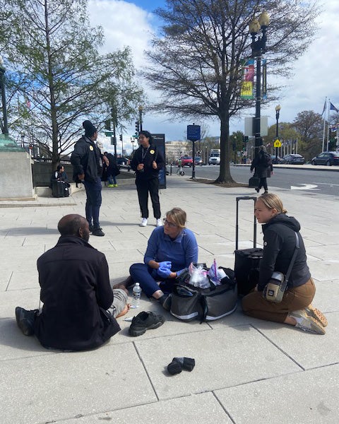 Several individuals listen as a person experiencing homelessness speaks to them on a sidewalk in an urban area