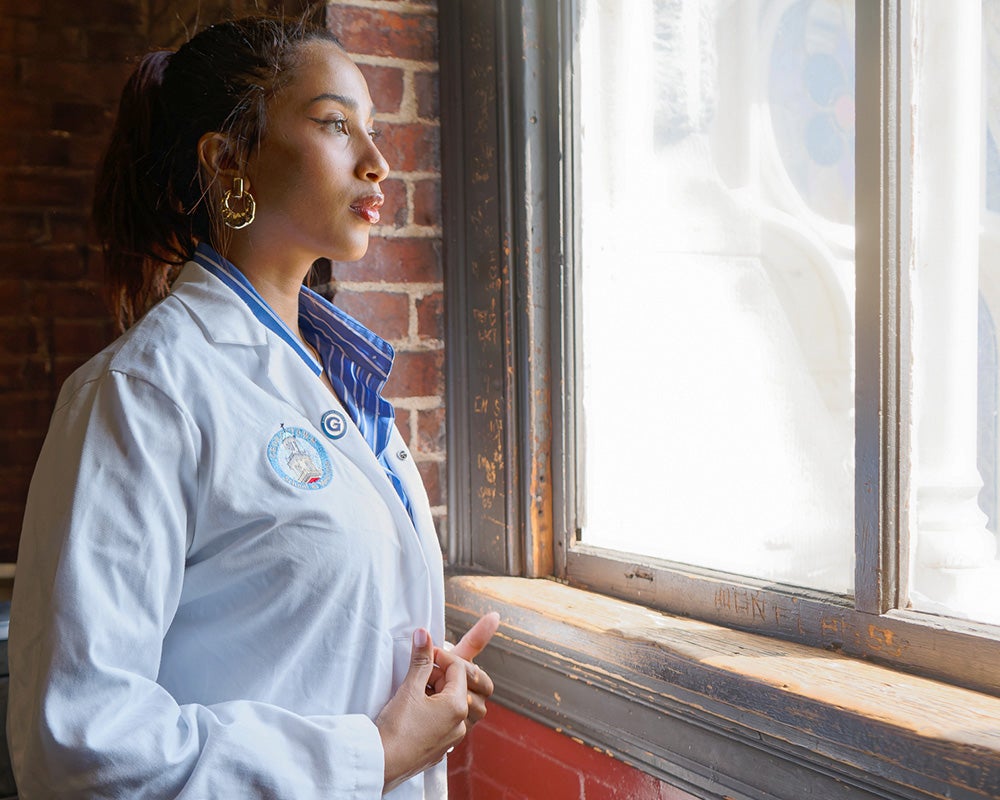 A medical student wearing a white coat looks out a window in Gaston Hall