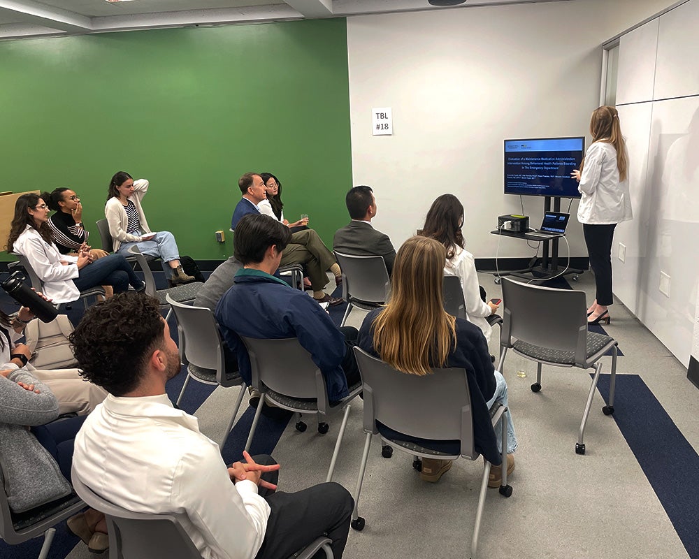 Students and faculty sit in chairs and listen to a student give her presentation