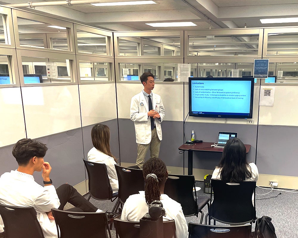 A student presents from next to a monitor displaying his presentation while other students sit in chairs before him