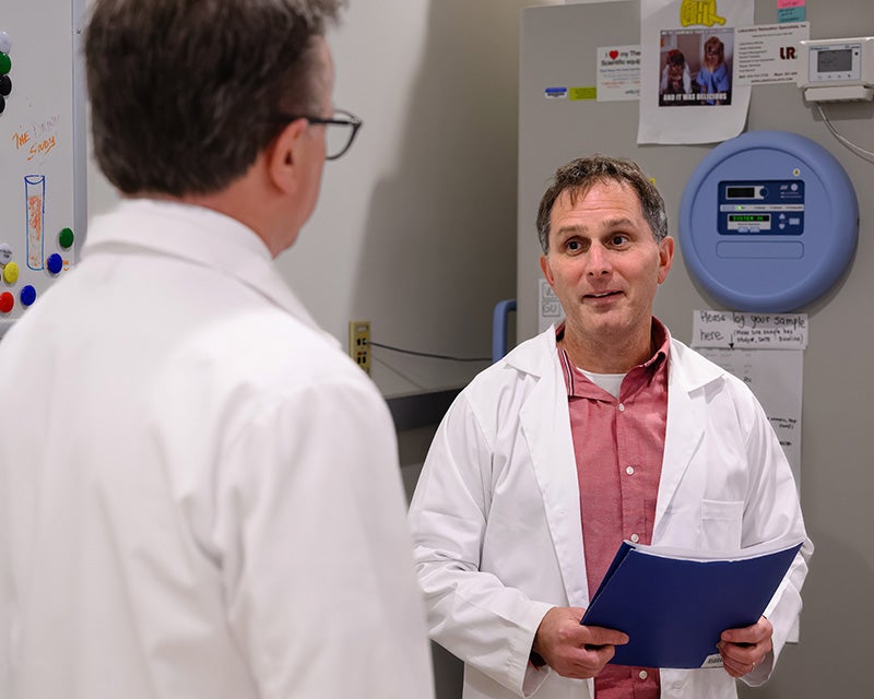 Dan Merestein stands in front of lab equipment and speaks with Dr. Beauchamp