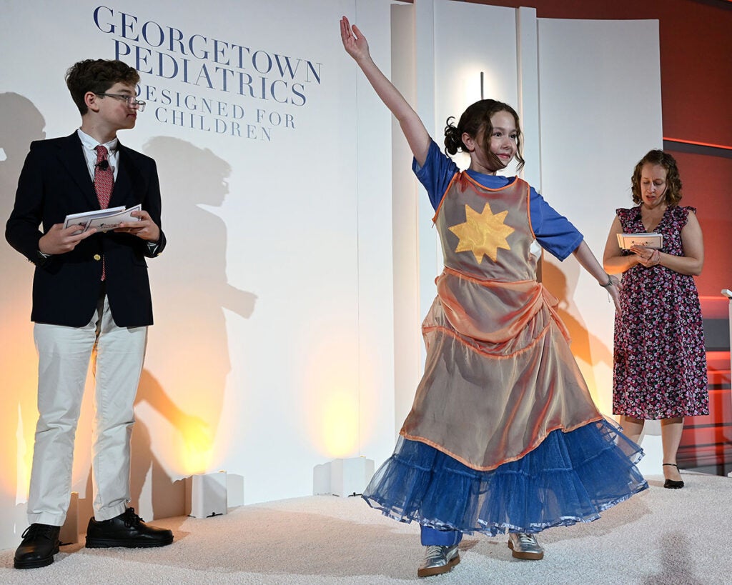 A young white girl holds out her arms to model a frilly dress emblazoned with a yellow star while the event emcees look on