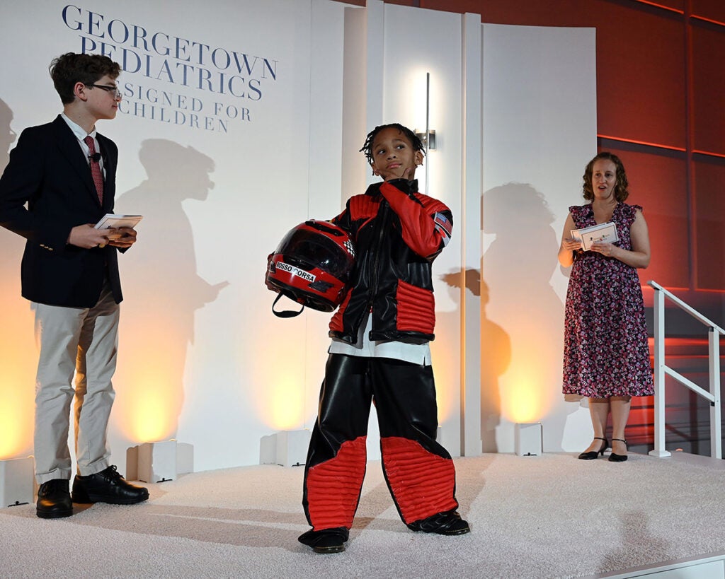 A young Black boy wears a racecar driver-like outfit of pants and jacket, holding a racing helmet in one arm while the event emcees stand behind him