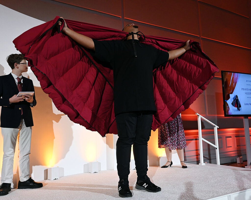 A young Black man holds his capelike garment in outstretched arms and looks at the ceiling