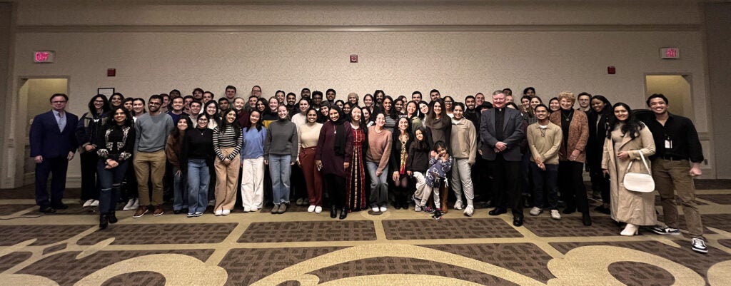 A large group of attendees at the Interfaith Dinner stands in the ballroom of the Leavey Center