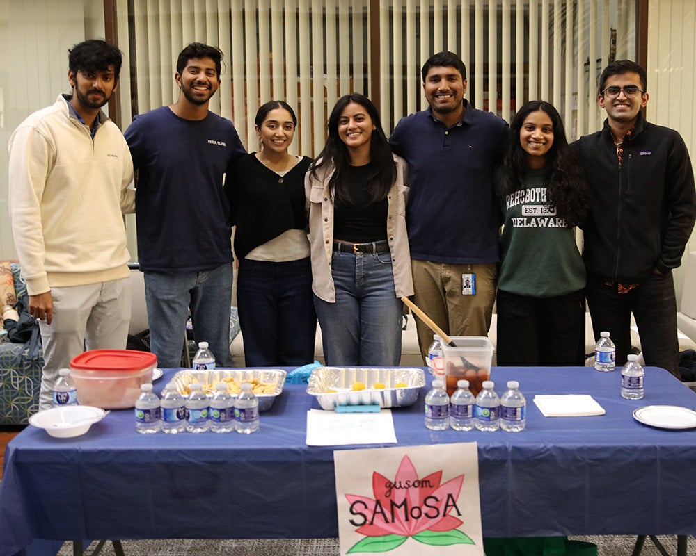 Seven students stand together behind a table with food items arrayed on it and a sign reading SAMOSA on the front