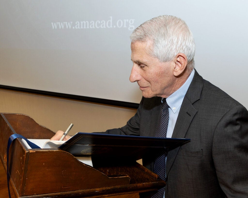 Anthony Fauci stands at a wooden podium and signs a book with a pen